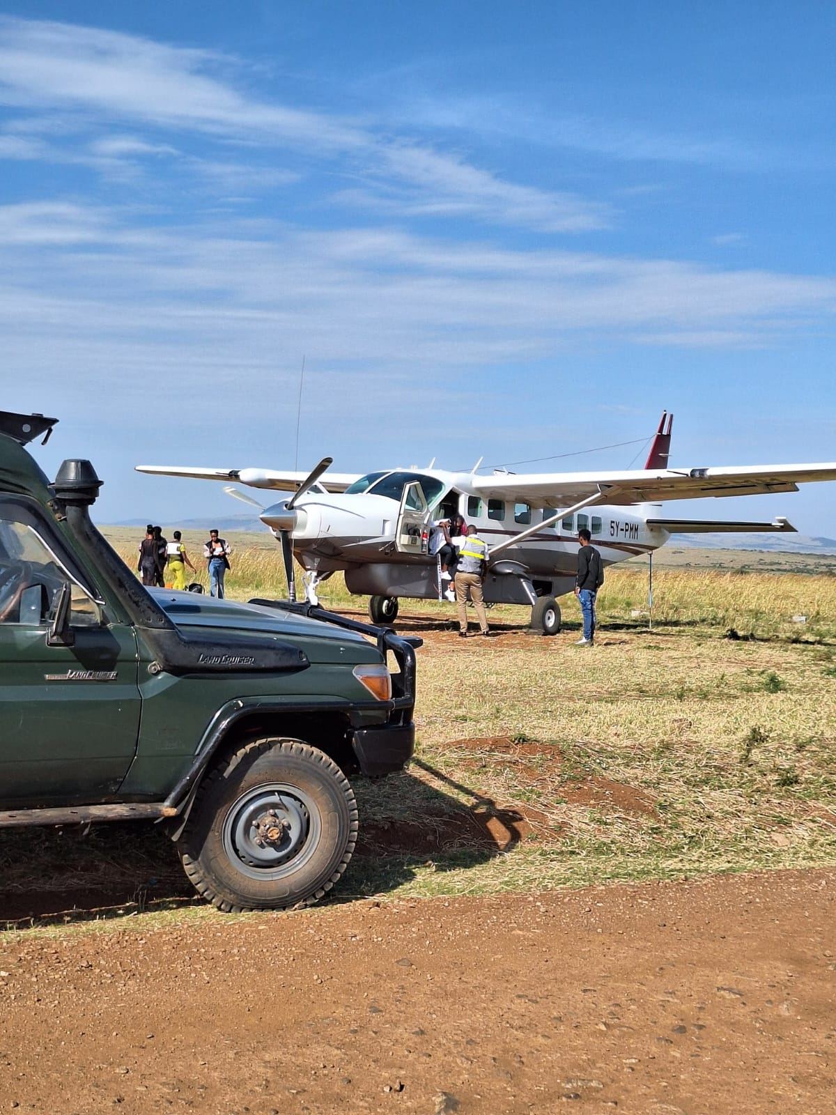 Cessna Grand Caravan 5Y-PMM boarding passengers on a grass airstrip beside a safari Land Cruiser